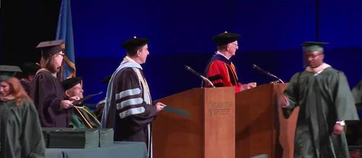 Graduation ceremony scene with students in caps and gowns walking on stage, speaker at budget encoder podium, and faculty seated in background.