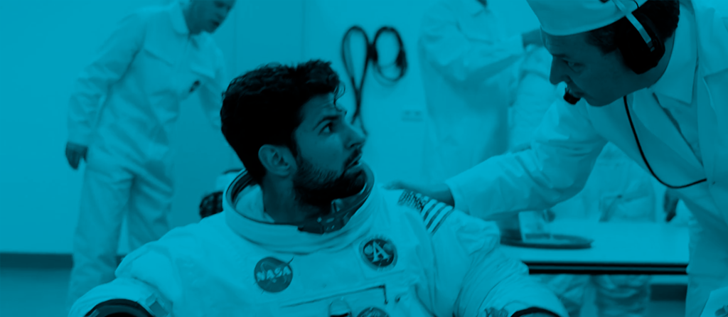 Astronaut, seated in a space suit, looks up at a technician speaking to him. Other personnel are visible in the background. The image is tinted blue.