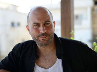 A bald man with a black shirt and white undershirt is standing outdoors.