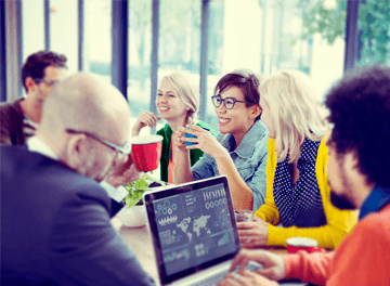 A group of people sit around a table engaged in conversation. One person has a laptop showing charts and graphs. Others are holding cups and appear to be enjoying a casual discussion.