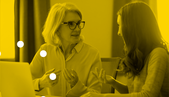 An older woman wearing glasses and a younger woman are seated at a table, engaged in conversation. A laptop is in front of them, and the image has a yellow tint with white dots.