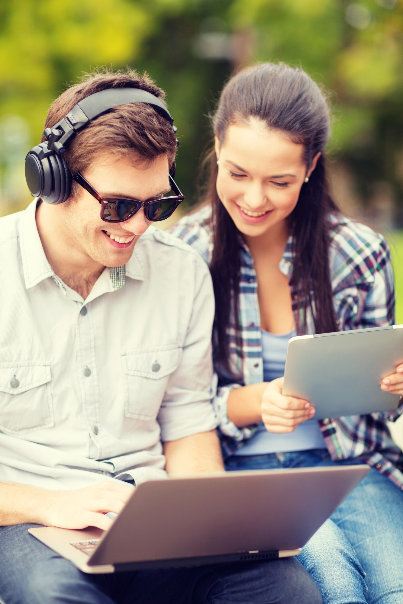 Two people sitting outdoors, with one wearing headphones and sunglasses using a laptop, and the other holding a tablet. Both appear to be looking at the laptop screen and smiling.
