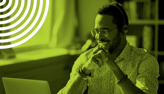 A man wearing glasses and headphones smiles while looking at a laptop screen in a brightly lit room.