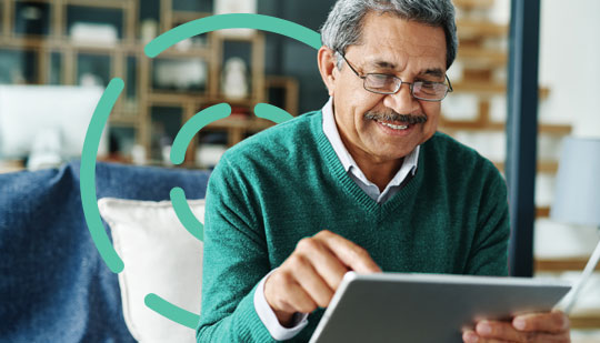 An older man with glasses is sitting indoors, using a tablet and smiling. He is wearing a green sweater and has a short mustache. Bookshelves are visible in the background.