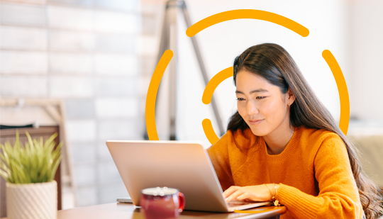 A person with long hair wearing an orange sweater is working on a laptop at a table. A mug and a small potted plant are also on the table.