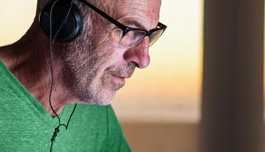 A man with glasses and a green shirt wearing headphones, looking intently at something off-frame.