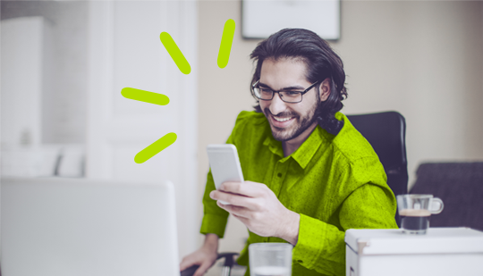 A man in a green shirt, seated at a desk in front of a laptop, smiles while looking at his smartphone. A small cup of coffee and a glass of water are on the desk.