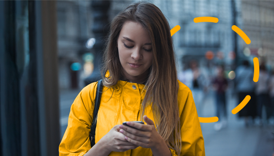 A woman in a yellow jacket stands on a city street, immersed in her smartphone, possibly streaming OTT content.
