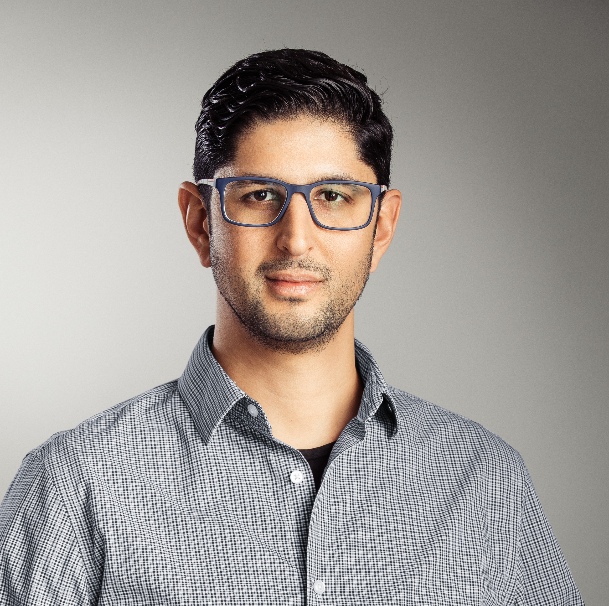 A man with short dark hair, wearing glasses and a checkered shirt, stands against a neutral gray background.