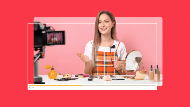 A person sits at a table with makeup products, smiling at a camera on a tripod. They are demonstrating makeup application against a pink background.