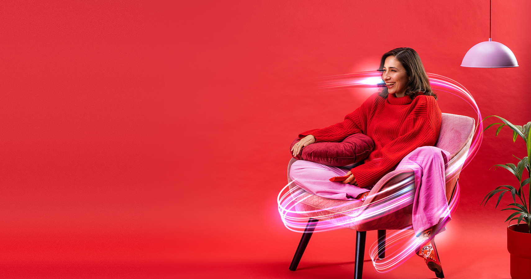 A woman in a red sweater and pink pants sits on a chair holding a pillow. Colorful light trails surround her, and a plant is next to her. The background is entirely red with a hanging white lamp.