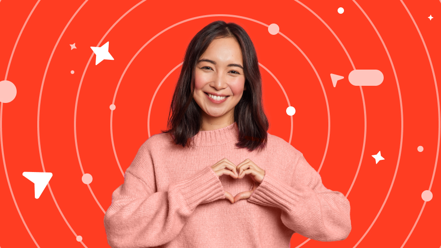 A smiling person with shoulder-length hair wears a pink sweater and makes a heart shape with their hands against a red background with star and circle patterns.