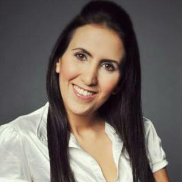 A woman with long dark hair is smiling at the camera. She is wearing a white blouse and standing against a dark grey background.