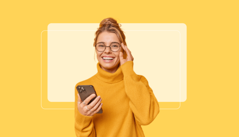 A person wearing glasses and a mustard yellow turtleneck sweater is holding a smartphone and smiling against a yellow background.